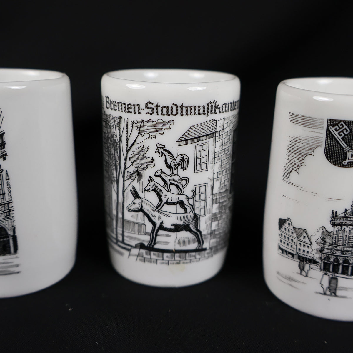 Detailed close-up of the Bremen Stadtmusikanten souvenir cup positioned between portions of the other two cups. The black illustration depicts a donkey, dog, cat, and rooster stacked together before buildings, with the German landmark title printed along the top.
