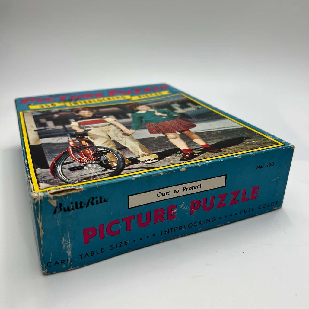 Angled view of the original Built-Rite picture puzzle box showing the Ours to Protect title label and No. 300 on the side. The lid depicts children beside a red bicycle, with blue packaging, pink lettering, yellow accents, and worn corners and edges.