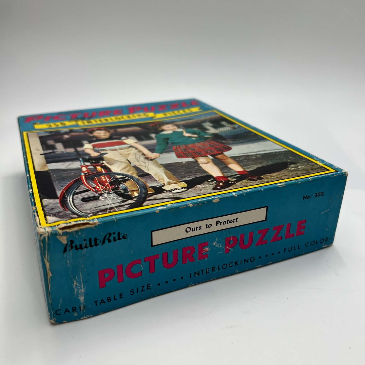 Angled view of the original Built-Rite picture puzzle box showing the Ours to Protect title label and No. 300 on the side. The lid depicts children beside a red bicycle, with blue packaging, pink lettering, yellow accents, and worn corners and edges.