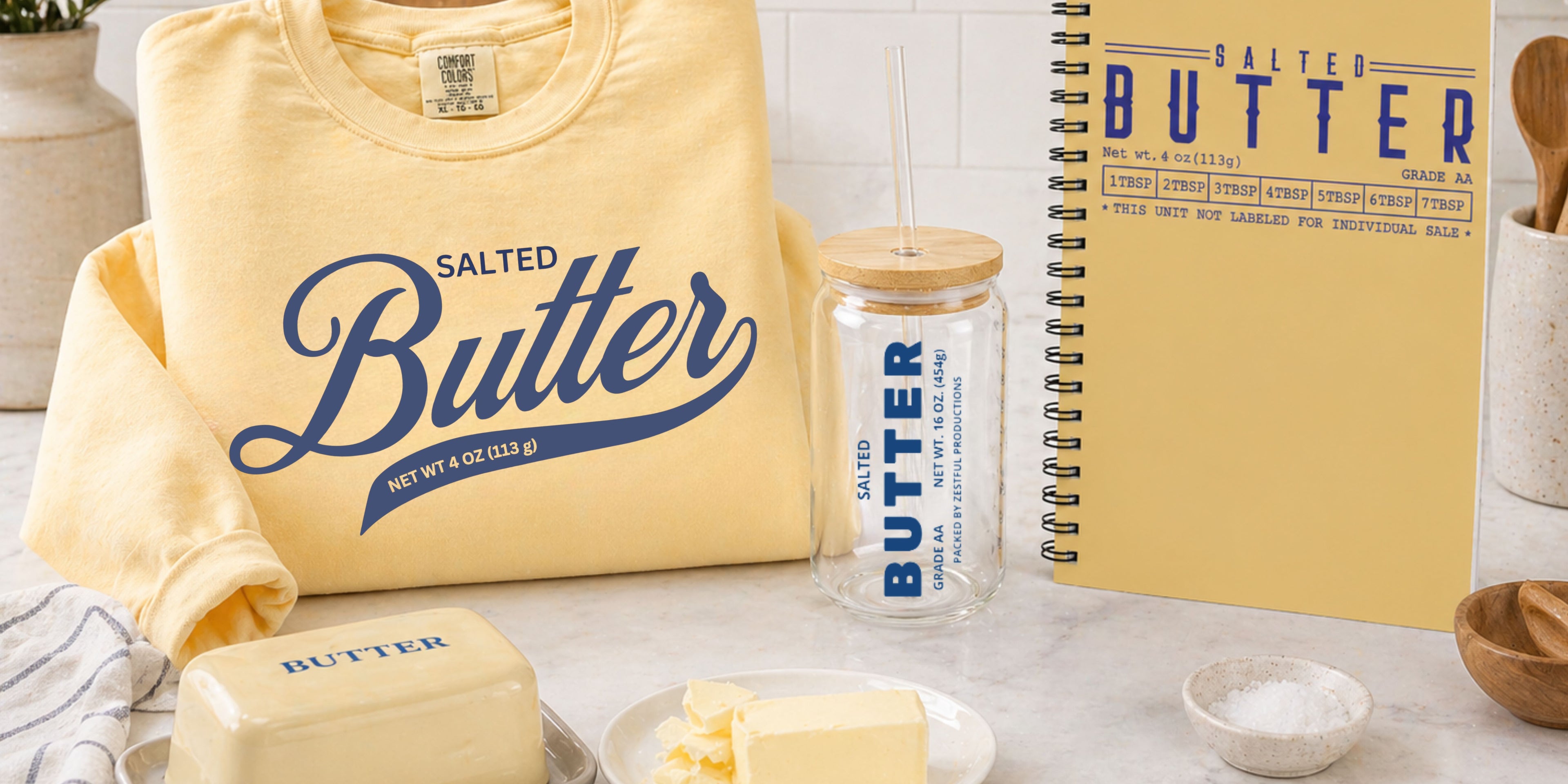 Modern kitchen lifestyle scene featuring Zestful Productions butter-themed products including a yellow “Salted Butter” sweatshirt, glass sipper jar, spiral notebook, butter dish, and fresh butter on marble countertop.