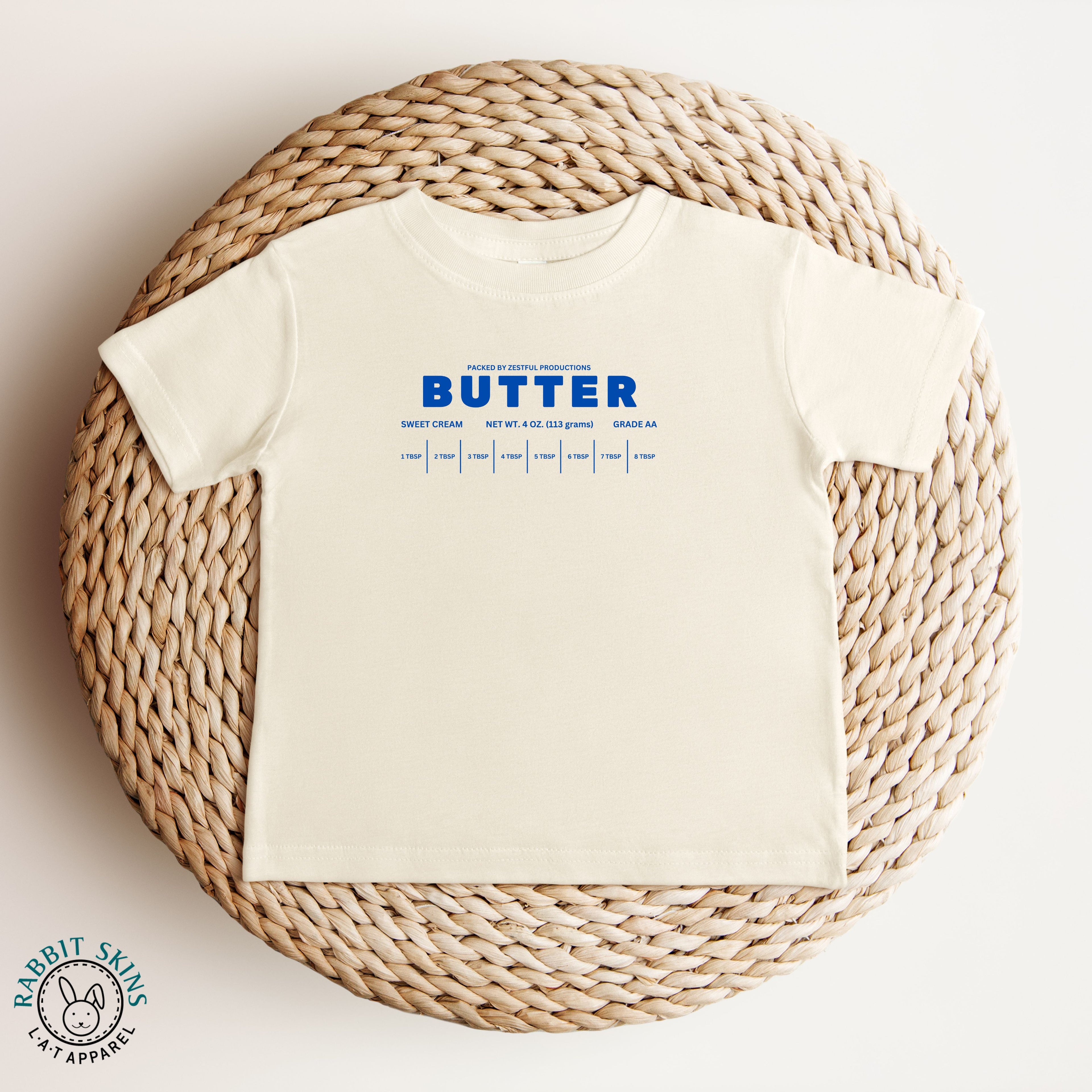 Natural color butter infant tee with bold blue lettering in flat lay. Neutral baby outfit piece with simple playful design.