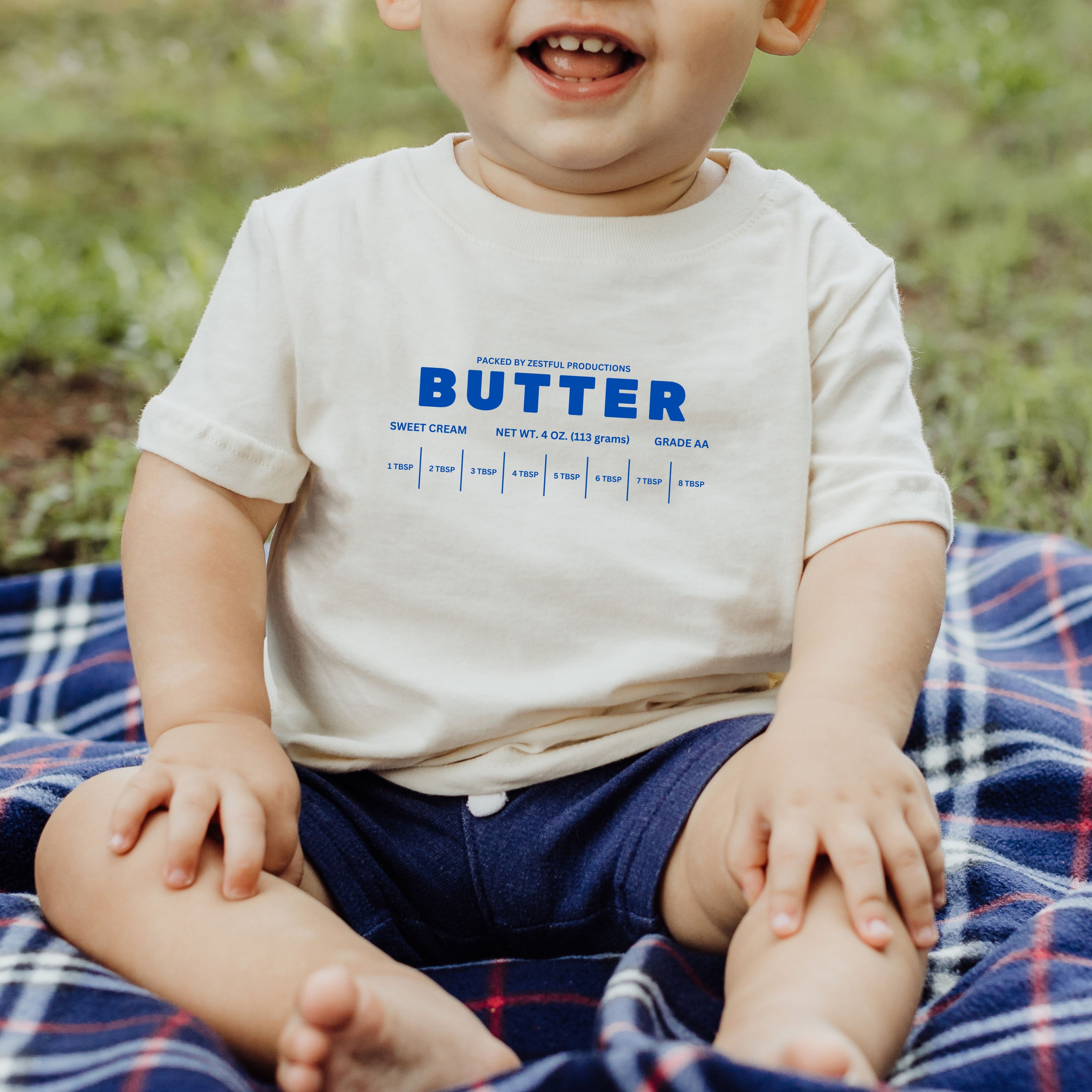 Baby wearing butter infant tee in soft cream color sitting outdoors. Cute baby outfit with bold butter graphic design, perfect for everyday wear, photos, and gifting.