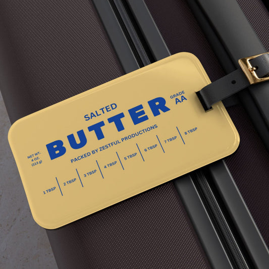 Butter luggage tag attached to suitcase with bold butter stick design and blue lettering. Funny travel accessory that makes bags easy to spot and adds personality to luggage.