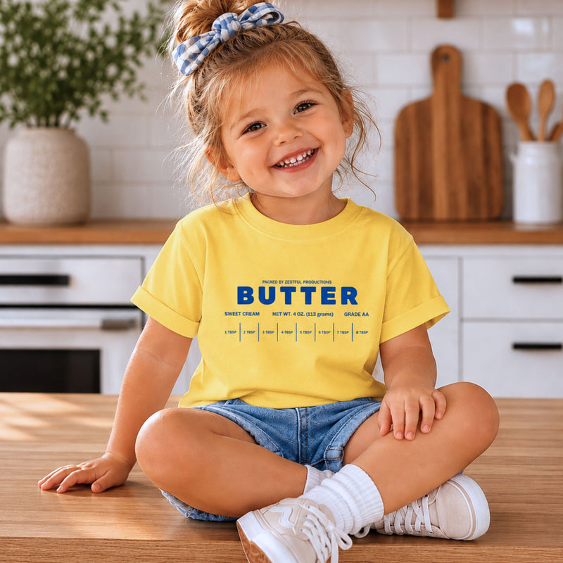Smiling toddler girl wearing yellow butter toddler tee with retro foodie graphic in boutique kitchen setting. Sweet cream inspired birthday outfit styled for preschool parties, playful everyday wear, and trendy kids fashion photography.