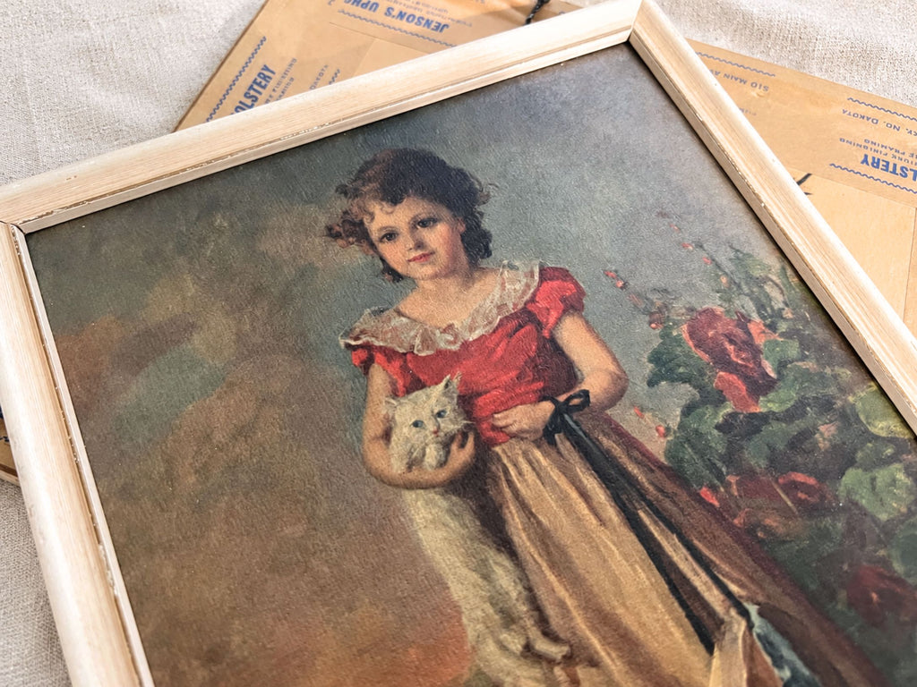 Close view of the girl-with-cat portrait, showing her dark curls, red bodice, pale ruffled collar, golden skirt, and white cat. The cream frame runs diagonally across the image, with part of the other picture’s brown backing and printed tape behind it.