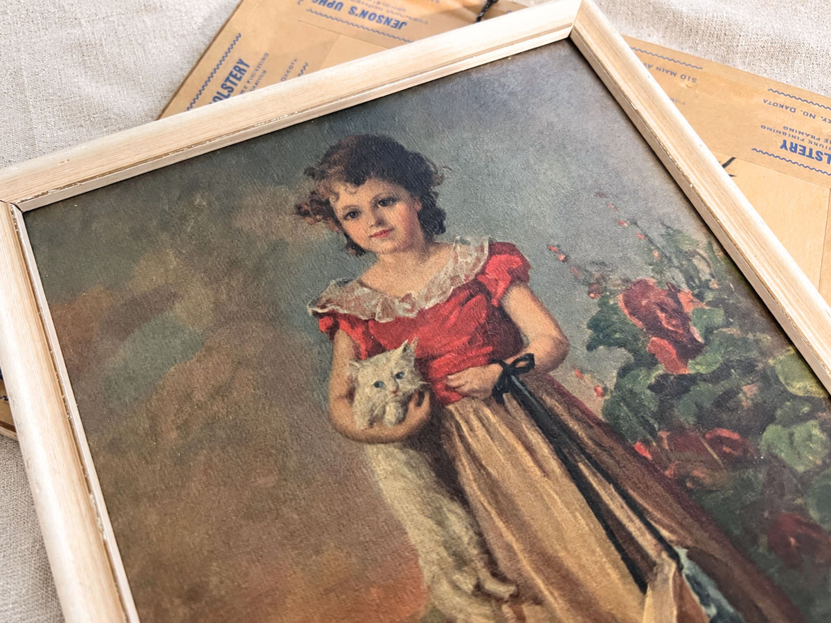 Close view of the girl-with-cat portrait, showing her dark curls, red bodice, pale ruffled collar, golden skirt, and white cat. The cream frame runs diagonally across the image, with part of the other picture’s brown backing and printed tape behind it.