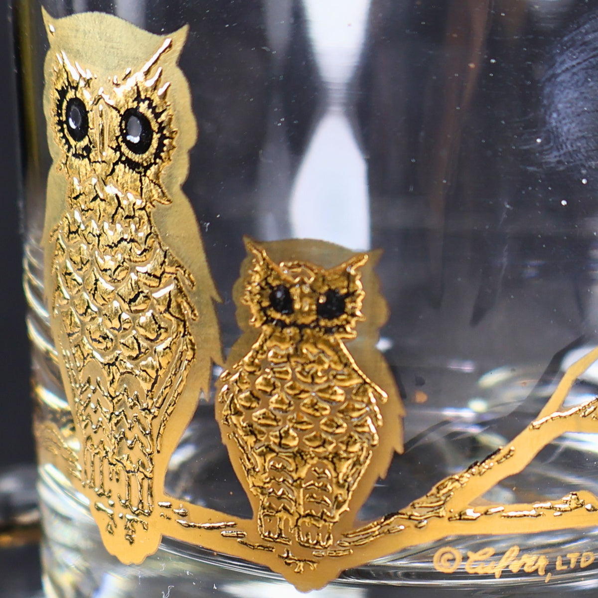 Macro photograph of the gold owl decoration on a vintage Culver rocks glass. Two owls with dark circular eyes sit on a gold branch, showing the raised feather texture, sculptural metallic finish, and fine decorative detail characteristic of Culver barware.