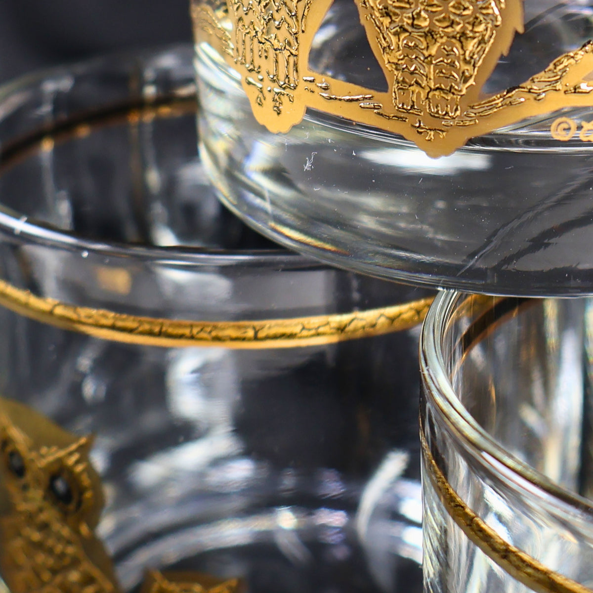 Condition detail of the vintage Culver owl glasses showing the gold rim bands, thick clear bases, and portions of the raised gold bird decoration. The close composition highlights the brightness of the metallic finish and the coordinated design across the set.