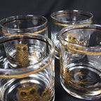 Four vintage clear Culver rocks glasses photographed closely from above and at an angle. Gold owl motifs appear through the transparent sides, while slim gold bands circle the rims and the heavy rounded bases catch reflections from the surrounding glassware.