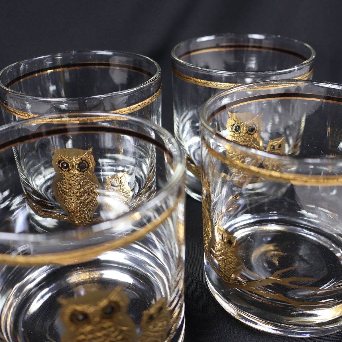 Four vintage clear Culver rocks glasses photographed closely from above and at an angle. Gold owl motifs appear through the transparent sides, while slim gold bands circle the rims and the heavy rounded bases catch reflections from the surrounding glassware.