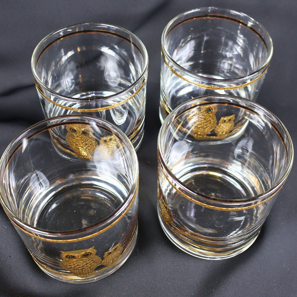 Overhead view of four clear Culver Ltd. cocktail glasses grouped together, revealing their round openings, substantial glass bases, and coordinating gold rim bands. Portions of the gold owl-and-branch designs are visible through the transparent glass walls.