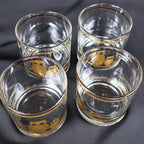 Overhead view of four clear Culver Ltd. cocktail glasses grouped together, revealing their round openings, substantial glass bases, and coordinating gold rim bands. Portions of the gold owl-and-branch designs are visible through the transparent glass walls.