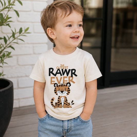 Smiling toddler wearing a beige Bella Canvas shirt with “Cutest RAWR Ever” tiger design, paired with denim shorts in a casual outdoor lifestyle setting.