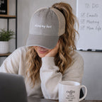 Woman wearing gray and tan mesh back trucker hat with doing my best embroidered saying while working on laptop with coffee mug