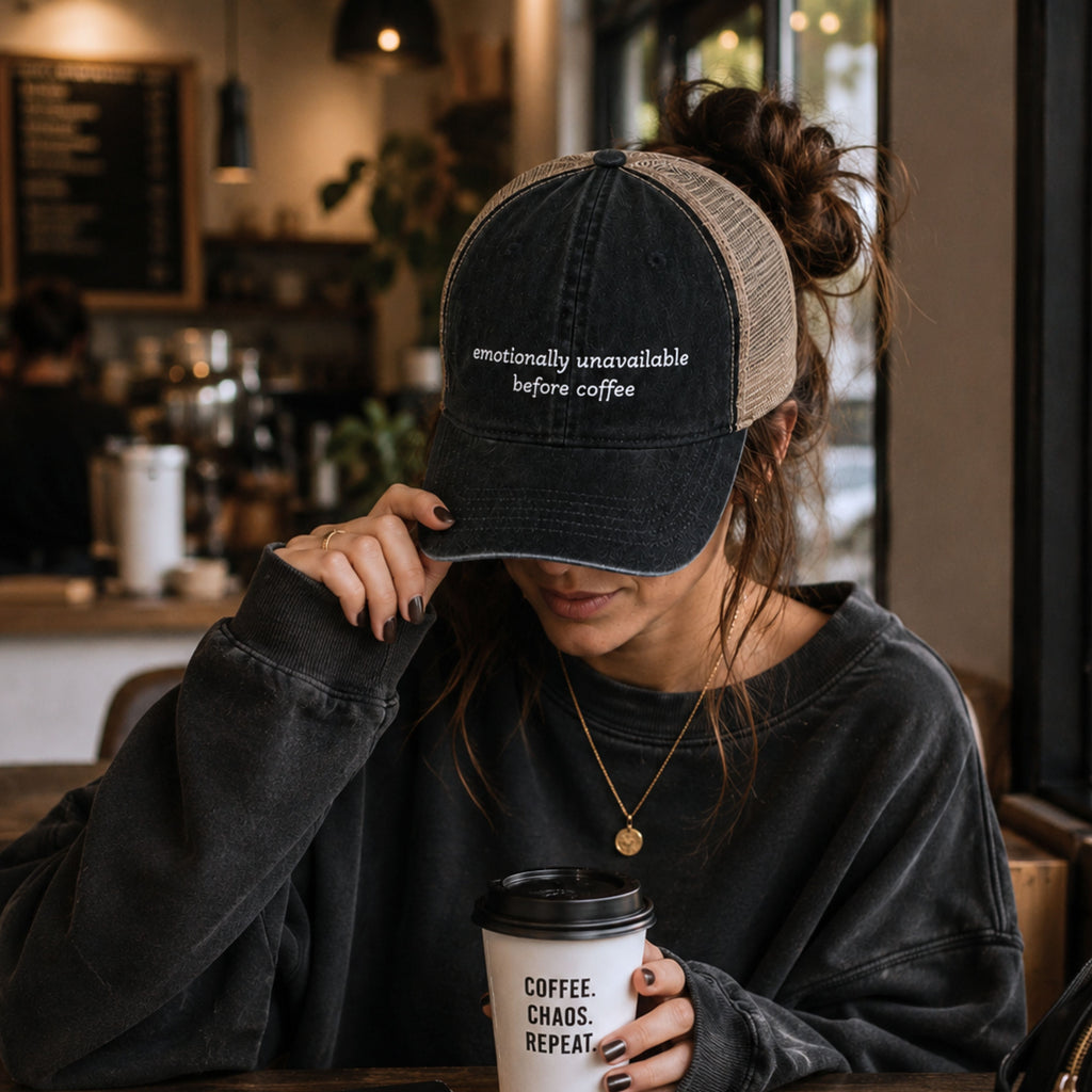 Woman wearing emotionally unavailable before coffee embroidered trucker hat while holding coffee in a cozy coffee shop