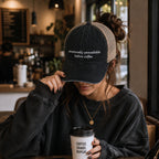 Woman wearing emotionally unavailable before coffee embroidered trucker hat while holding coffee in a cozy coffee shop