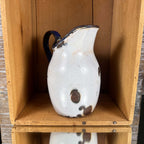 Opposite side of a white enamelware duckbill pitcher, with its curved blue handle on the left and raised spout on the right. Scratches and scattered dark coating losses mark the body, with chipped enamel and brown discoloration around the rim and handle attachments.