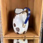 Rear angled view of a small white enamelware jug showing its cobalt-blue loop handle and two attachment points. Chipped enamel, brown rust, and discoloration surround the attachments, while larger dark patches appear on the body against the wooden crate backdrop.