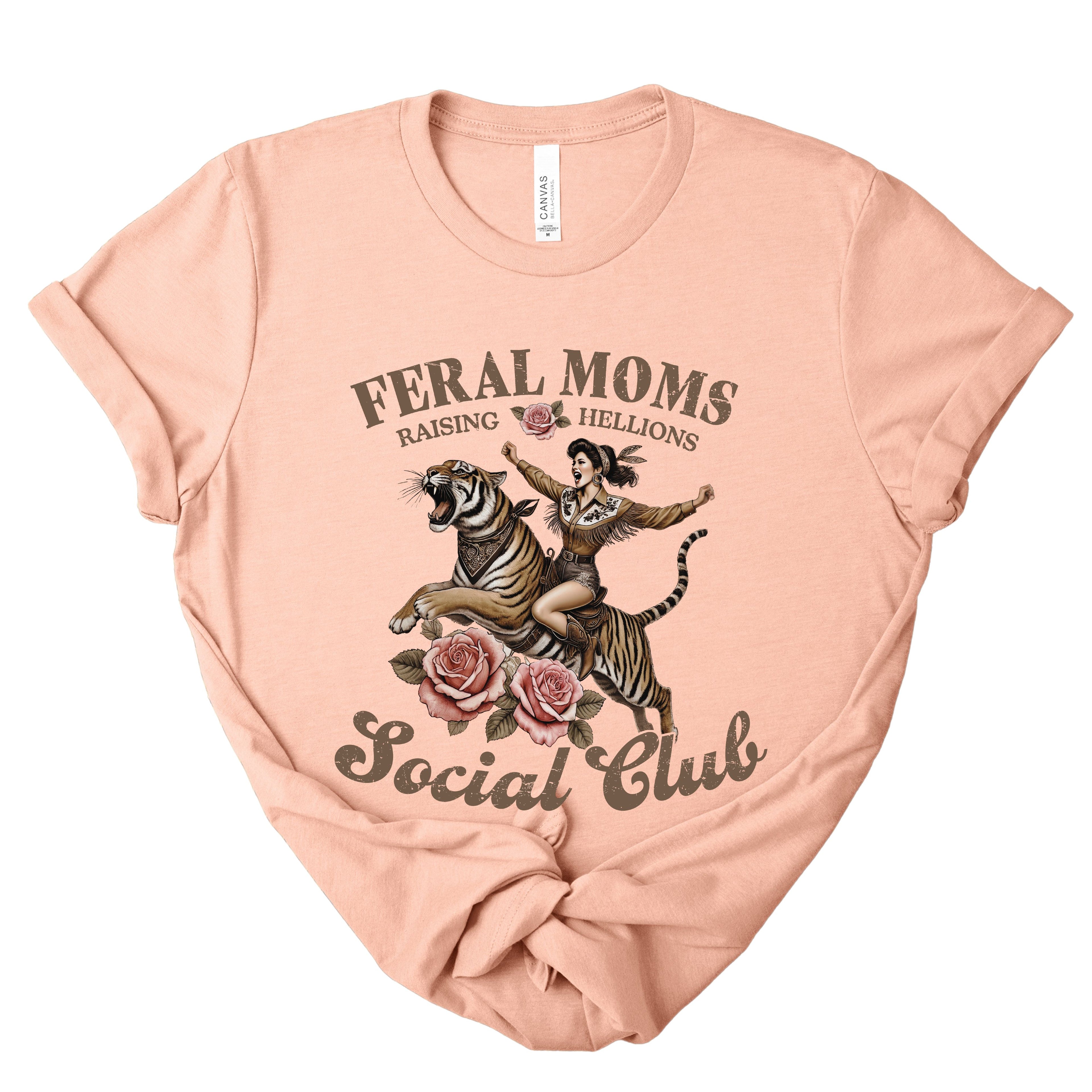 Heather peach t-shirt with graphic design of a woman riding a tiger and text 'Feral Moms Raising Hellions Social Club' on a white background.