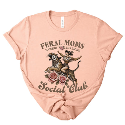 Heather peach t-shirt with graphic design of a woman riding a tiger and text 'Feral Moms Raising Hellions Social Club' on a white background.