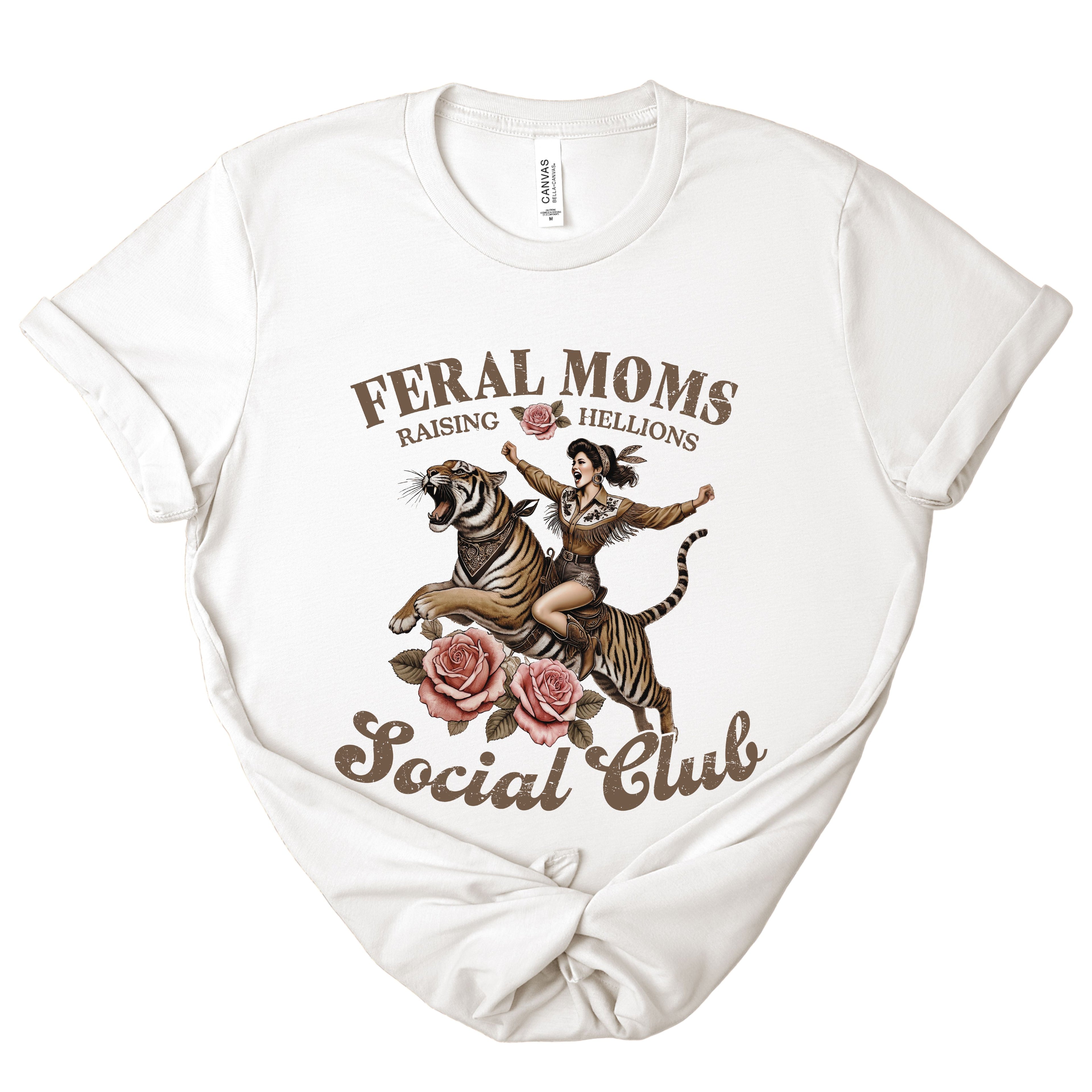 White t-shirt with graphic design of a woman riding a tiger and text 'Feral Moms Raising Hellions Social Club' on a white background.