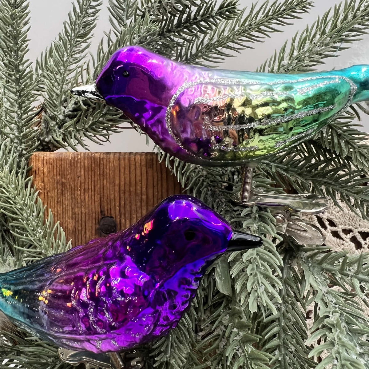 Close view of two glass clip-on bird ornaments nestled in evergreen branches. Purple heads, molded feather details, and silver-glitter wing outlines are visible, with green coloring on the upper bird and a violet body on the lower bird against wood and cream lace.
