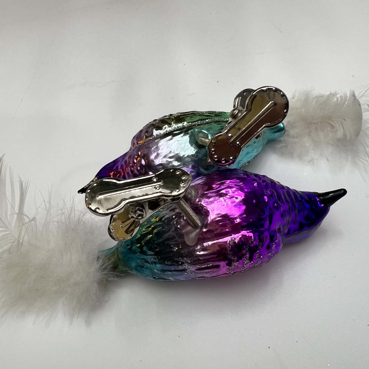 Underside view of two purple and teal glass bird ornaments lying on their sides on a white surface. The photograph shows the metal branch clips, coiled spring supports, and attachment points beneath the reflective bodies, with white feather tails extending outward.