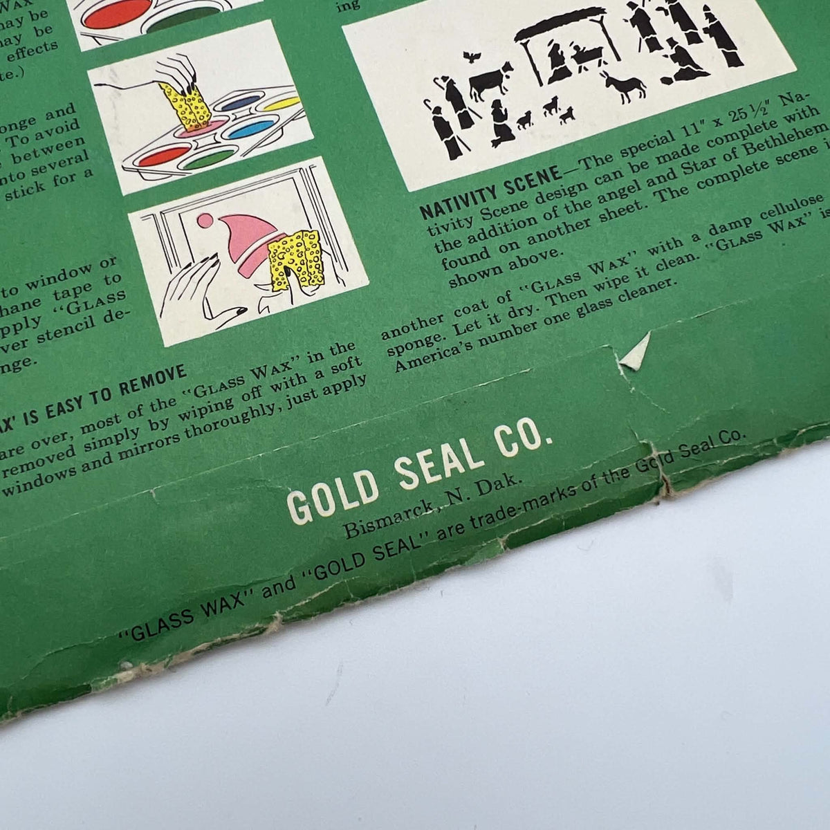 Close view of the green Glass Wax Christmas stencil envelope showing the Gold Seal Company name and Bismarck, North Dakota address. Nativity artwork and sponge application instructions appear above the label, with creases, edge wear, and a small tear visible.