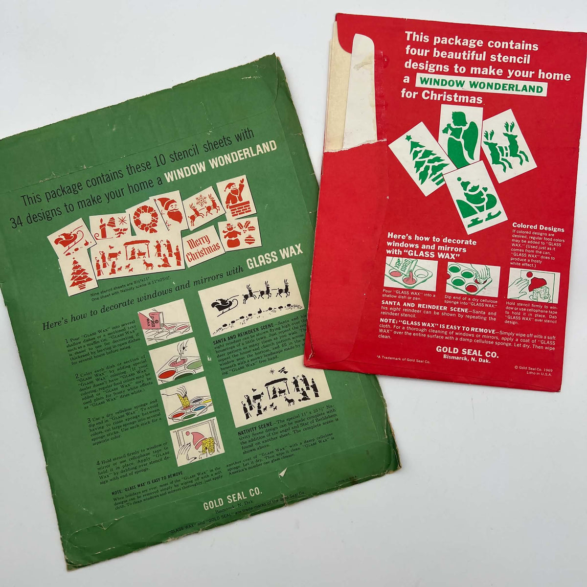 Reverse sides of green and red vintage Glass Wax Christmas stencil packages, showing illustrated decorating instructions, holiday design examples, and Gold Seal Company Bismarck labels. Creases, worn edges, and paper loss are visible on the original envelopes.