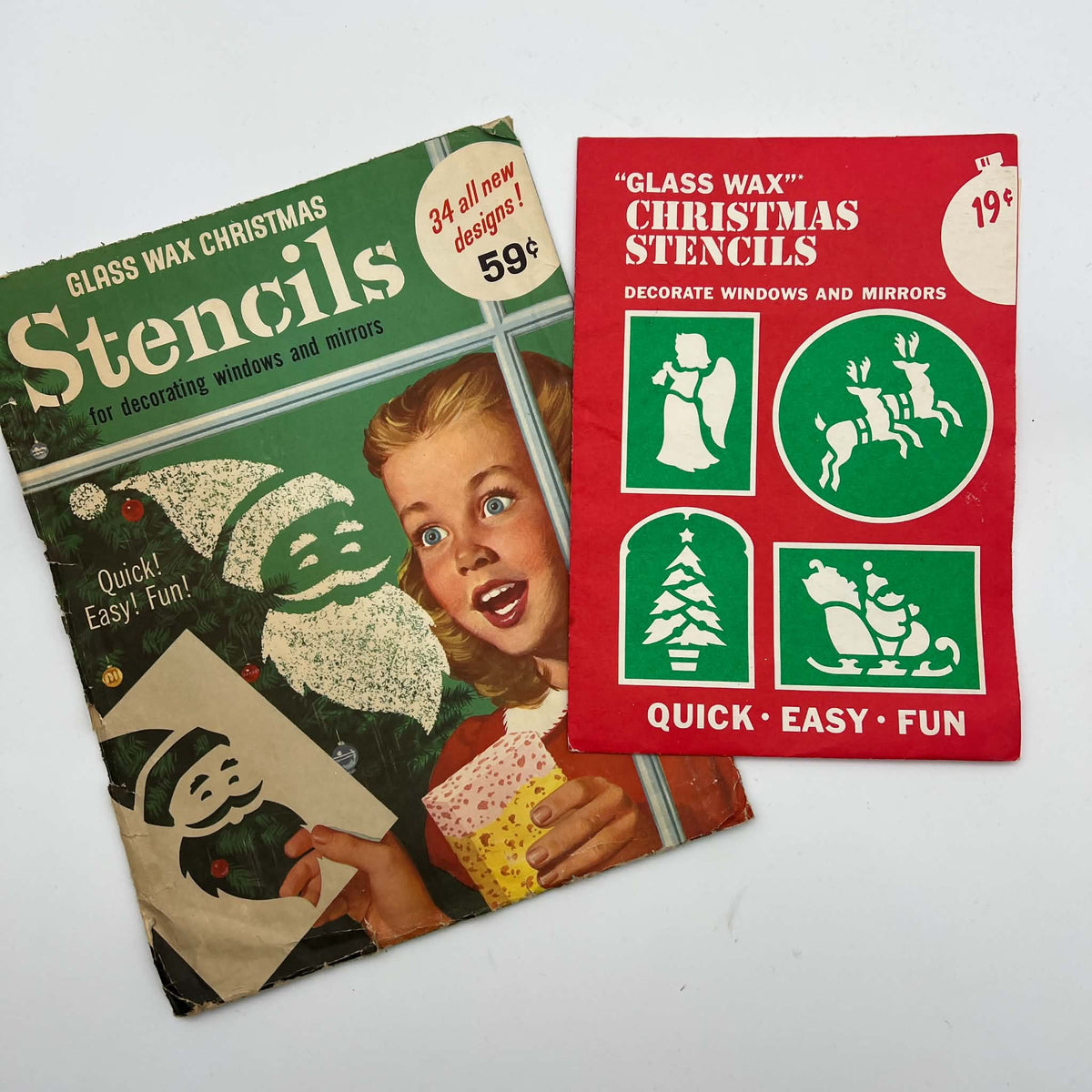 Two vintage Gold Seal Glass Wax Christmas stencil packages on a white surface. The larger green envelope shows a girl decorating a window with a Santa stencil; the red envelope displays angel, reindeer, tree, and sleigh designs with original printed prices.