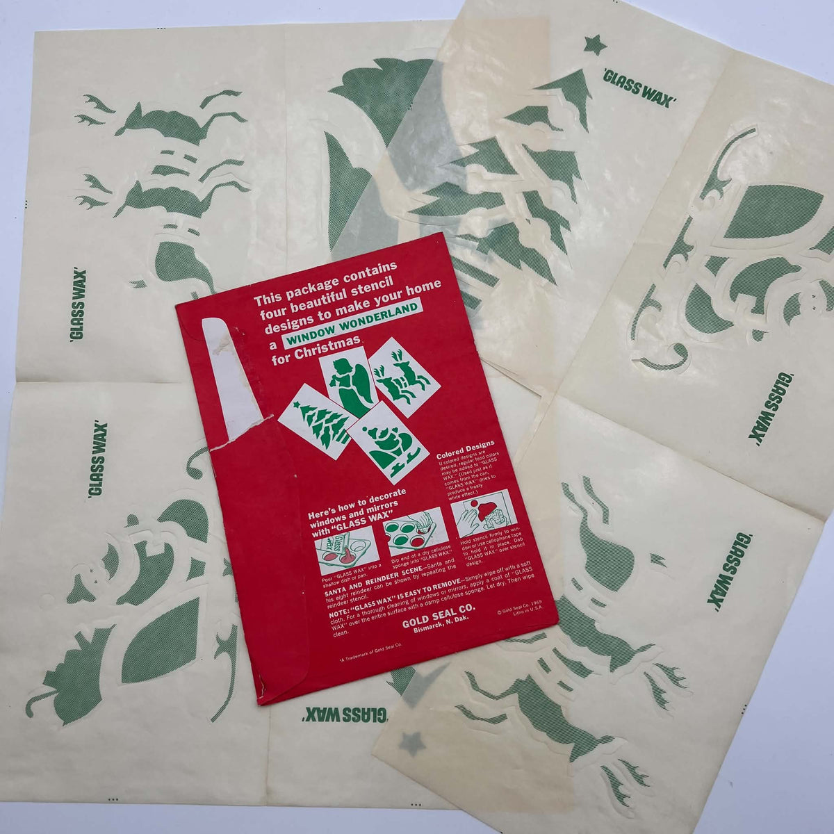 Four vintage Glass Wax Christmas stencil sheets arranged beneath their red Gold Seal envelope. Reindeer, Christmas tree, angel, and Santa sleigh motifs appear across cream paper with green printing and visible folds; the envelope shows instructions and paper loss.