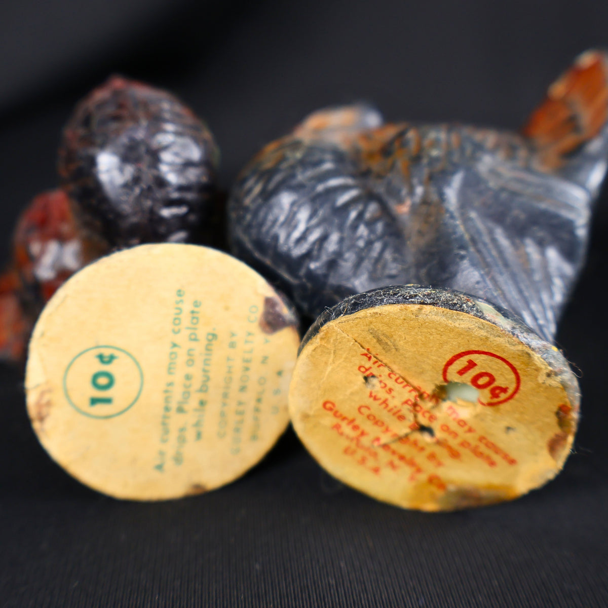 Underside view of two vintage Gurley Novelty Company turkey candles resting on their sides. Both circular paper labels display an original 10-cent price and printed candle-burning warning, while label staining, holes, edge wear and discoloration are also visible.