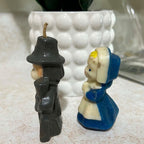 Side view of vintage Gurley pilgrim candles showing the male Thanksgiving figure's dark molded hat, clothing and long gun beside the female pilgrim's blue dress, cream apron and bonnet. Surface scuffs, discoloration and the dimensional wax construction are visible.
