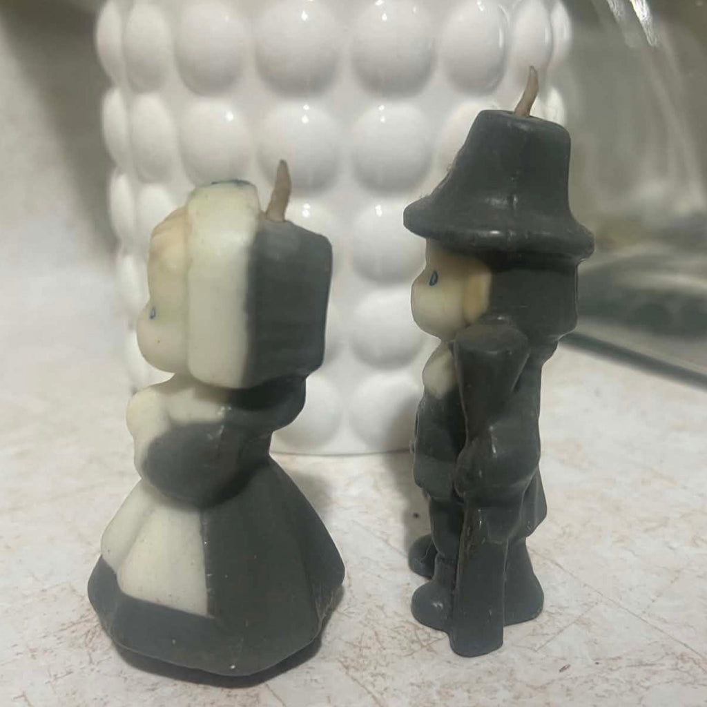 Opposite side profile of vintage Gurley pilgrim man and woman candles with their molded facial profiles, clothing and intact wicks visible. The photograph shows the cream and charcoal wax coloring along with the male pilgrim's hat, coat and long gun detail.