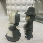 Opposite side profile of vintage Gurley pilgrim man and woman candles with their molded facial profiles, clothing and intact wicks visible. The photograph shows the cream and charcoal wax coloring along with the male pilgrim's hat, coat and long gun detail.