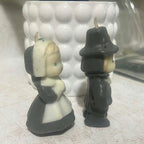 Side view of two vintage Gurley pilgrim candles showing the dimensional molded wax construction of the Thanksgiving figures. The woman wears a long cream and charcoal dress and bonnet while the male pilgrim has a broad dark hat, coat and long gun along his side.