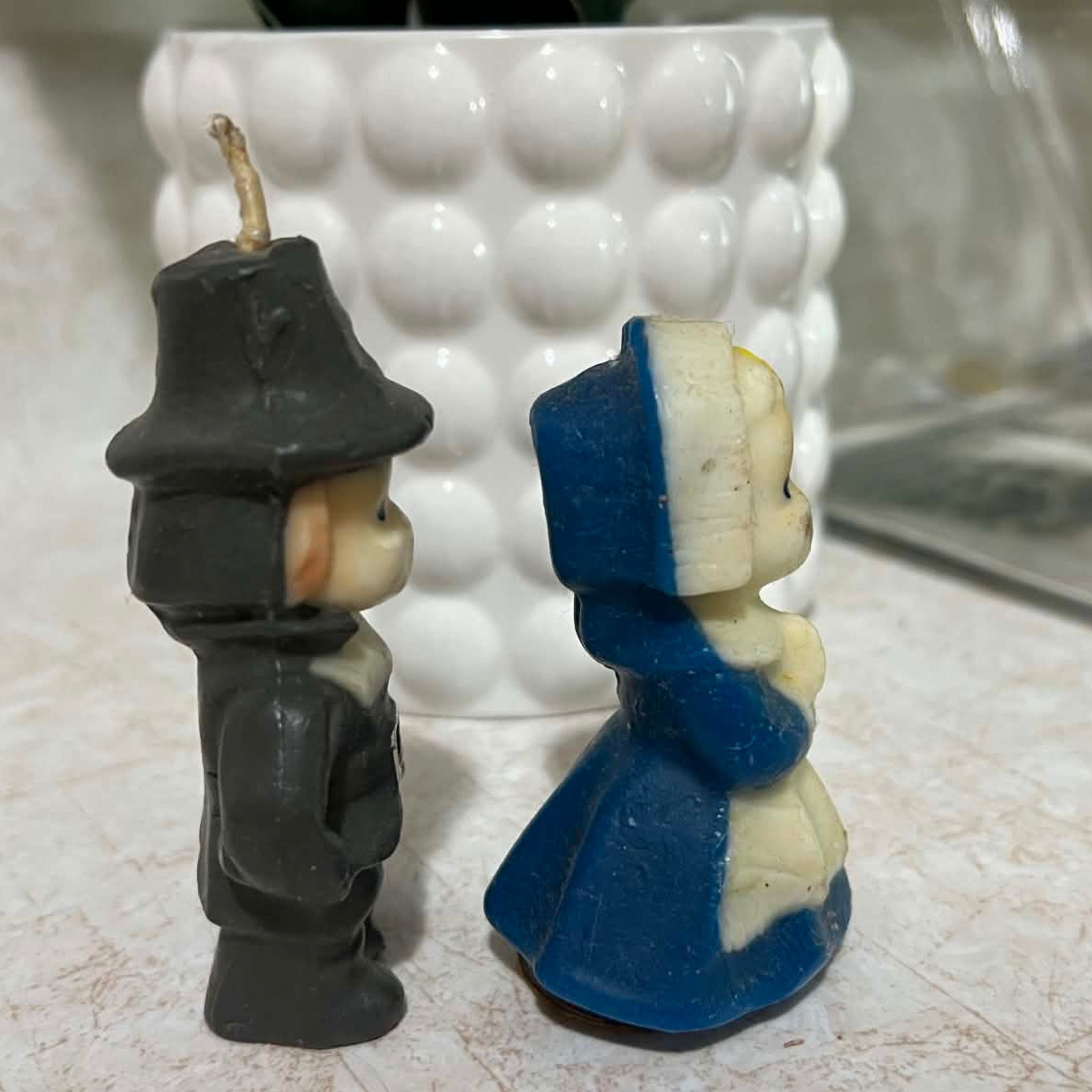 Opposite profile of vintage Gurley pilgrim candles with the male figure's long unburned wick, molded hat and dark clothing beside the female pilgrim's cream bonnet, clasped hands and blue dress. The photograph documents scratches, discoloration and wax wear.