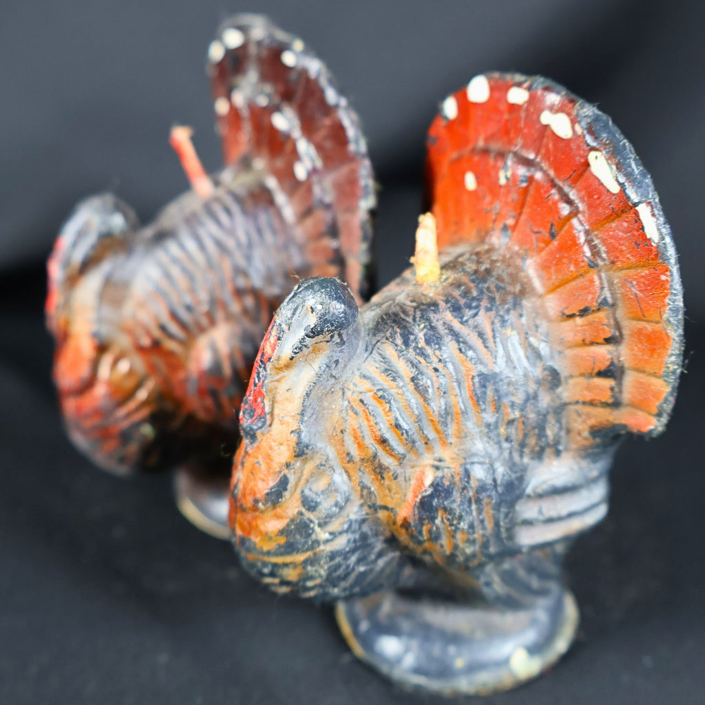 Angled close-up of two vintage Gurley Novelty Company turkey candles, with one wax figure in the foreground and the second behind it. Molded feather lines, orange and black coloring, red facial details, worn painted surfaces and unburned projecting wicks are visible.