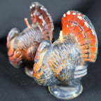 Angled close-up of two vintage Gurley Novelty Company turkey candles, with one wax figure in the foreground and the second behind it. Molded feather lines, orange and black coloring, red facial details, worn painted surfaces and unburned projecting wicks are visible.