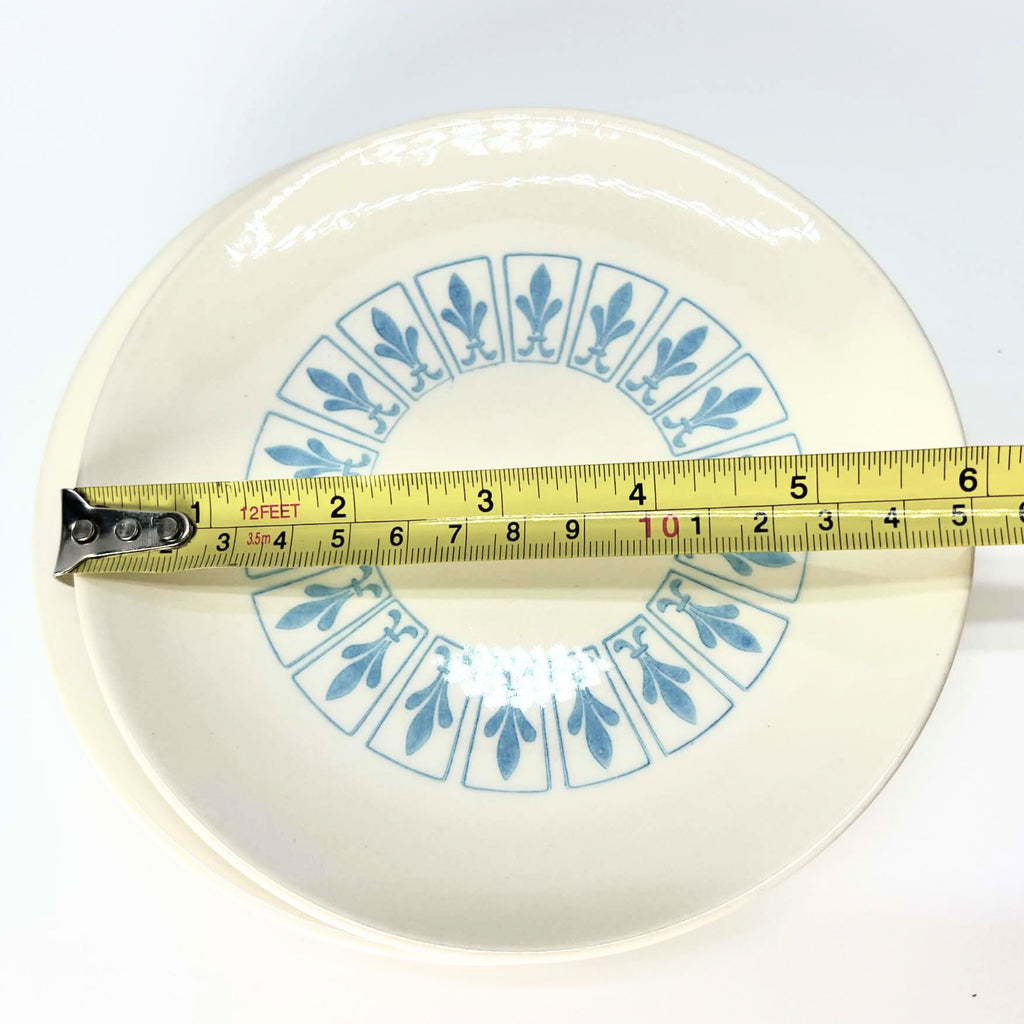 Ivory Richelieu bread-and-butter plate with blue fleur-de-lis motifs arranged in a circular band of outlined panels. A yellow tape measure stretches across the face, indicating an approximate diameter of six and a quarter inches against a white background.