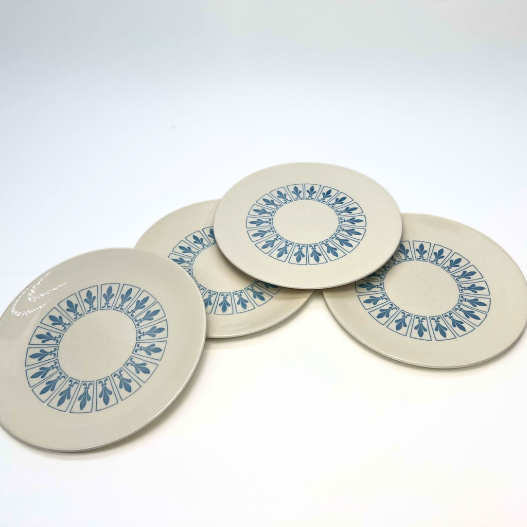 Four vintage Homer Laughlin Richelieu plates overlap in a loose fan arrangement on white. Each has a warm ivory surface with a circular band of blue fleur-de-lis panels, an undecorated center, and a broad plain outer margin around the patterned area.