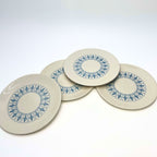Four vintage Homer Laughlin Richelieu plates overlap in a loose fan arrangement on white. Each has a warm ivory surface with a circular band of blue fleur-de-lis panels, an undecorated center, and a broad plain outer margin around the patterned area.