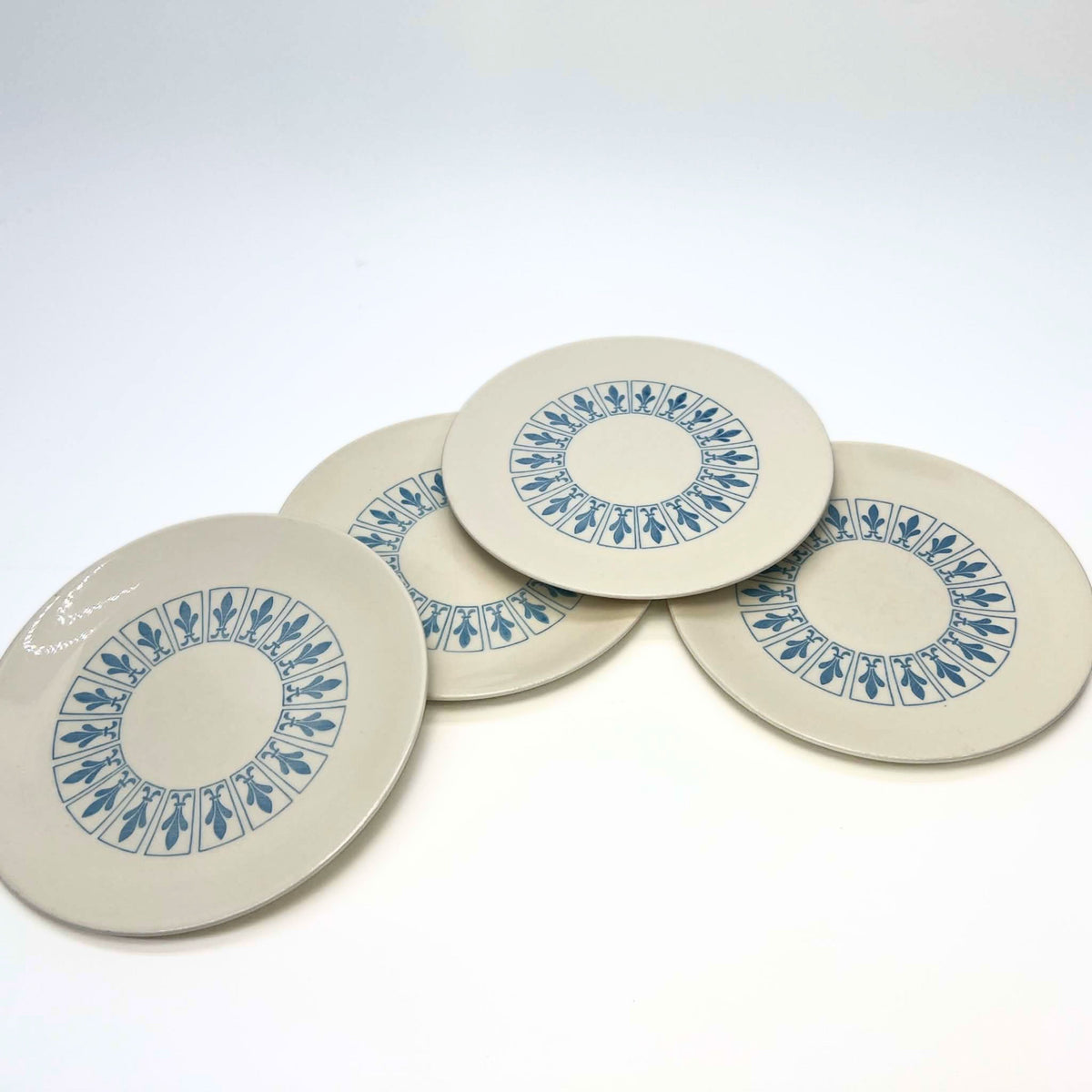Four vintage Homer Laughlin Richelieu plates overlap in a loose fan arrangement on white. Each has a warm ivory surface with a circular band of blue fleur-de-lis panels, an undecorated center, and a broad plain outer margin around the patterned area.
