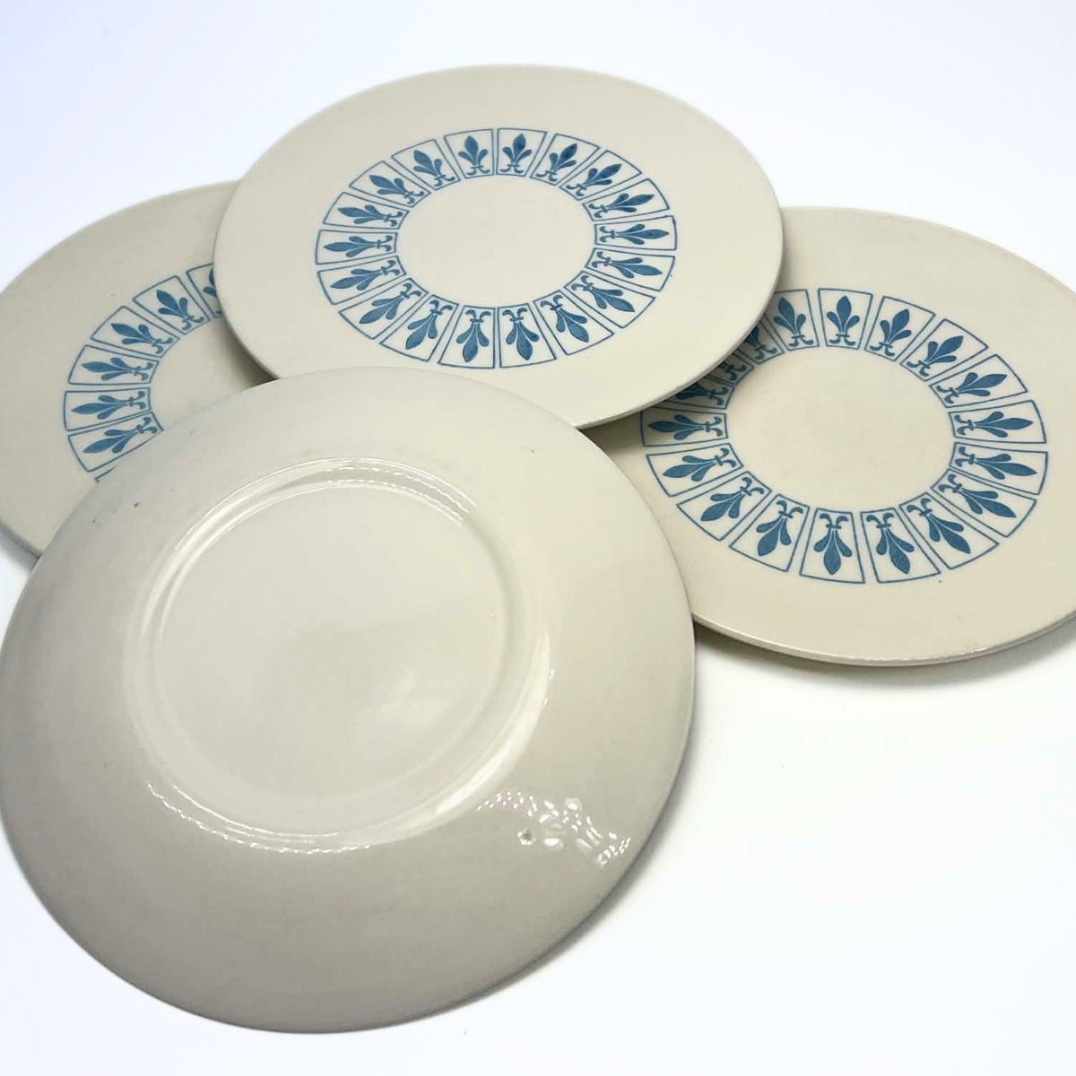 Four coordinating Richelieu plates arranged with three patterned faces visible behind one upturned plate. The fronts show blue fleur-de-lis panels on ivory, while the foreground plate reveals its plain glazed underside and raised circular foot ring.