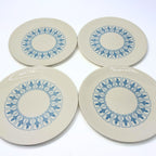 Set of four ivory bread-and-butter plates photographed face up in two rows. Blue fleur-de-lis emblems repeat inside outlined panels around each plain center, with smooth untrimmed edges and light reflecting across the glazed surfaces of the front plates.