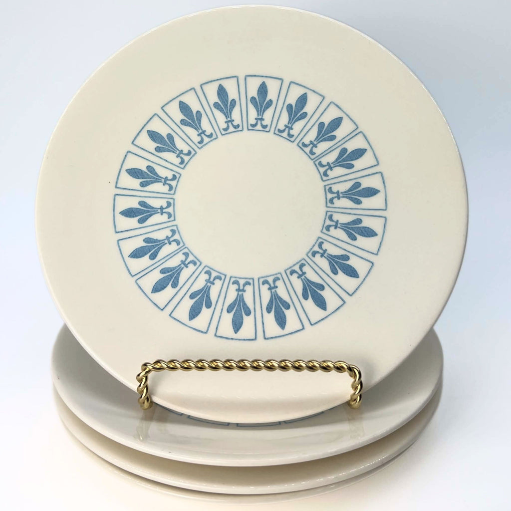One ivory Homer Laughlin Richelieu plate stands upright on a gold-colored twisted-wire stand above three stacked plates. Its blue fleur-de-lis panel design forms a complete ring around the plain center, with the smooth outer edge clearly visible.