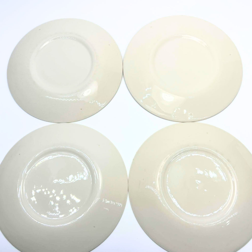 Undersides of four cream-colored plates arranged in two rows on a white background. Each has a circular foot ring and glossy glaze with reflected light. Small scattered specks are visible, and no printed maker’s marks appear on the exposed bases.