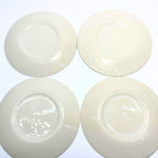 Undersides of four cream-colored plates arranged in two rows on a white background. Each has a circular foot ring and glossy glaze with reflected light. Small scattered specks are visible, and no printed maker’s marks appear on the exposed bases.