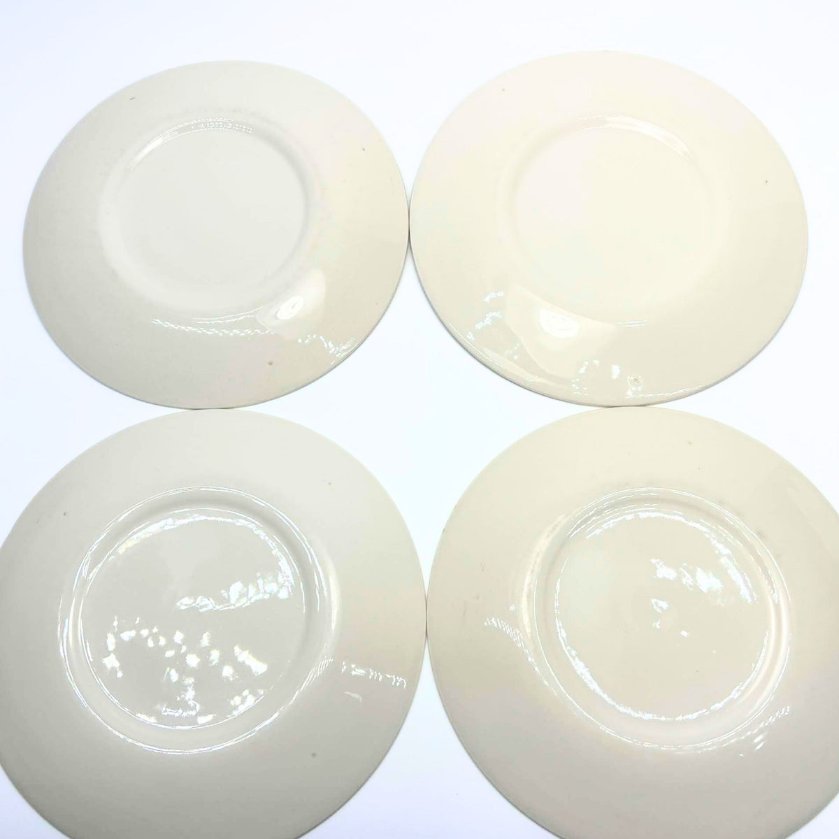 Undersides of four cream-colored plates arranged in two rows on a white background. Each has a circular foot ring and glossy glaze with reflected light. Small scattered specks are visible, and no printed maker’s marks appear on the exposed bases.