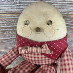 Front portrait of a Humpty Dumpty cloth doll showing button eyes, stitched circular cheeks, a rounded brown nose and a short stitched mouth. Small blue-green marks appear on the cream face above a red heart-patterned collar, striped bow and checked outfit.